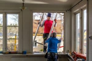 A renovation project in progress as two workers replace an old window. One man passes the frame from the outside while the other prepares the opening from inside the home.