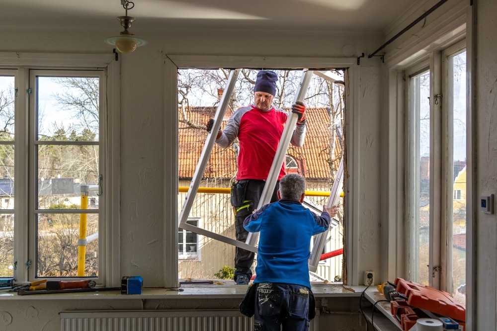 A renovation project in progress as two workers replace an old window. One man passes the frame from the outside while the other prepares the opening from inside the home.